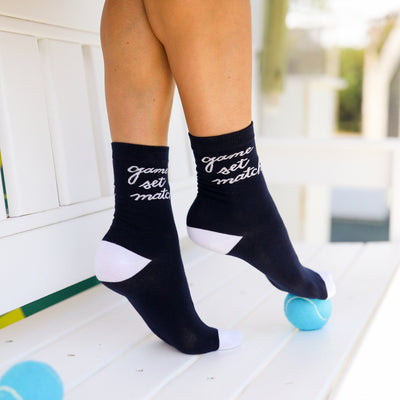 woman sits on a bench next to tennis court wearing a pair of navy socks with white heel and toes. White text that reads game set match in cursive font around ankle