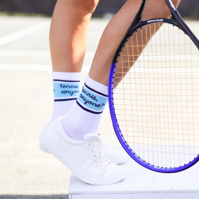 Woman stands on tennis court wearing a pair of white socks with navy heel and blue toes. Navy and blue stripes wrap around ankle of sock with the text tennis anyone in cursive font