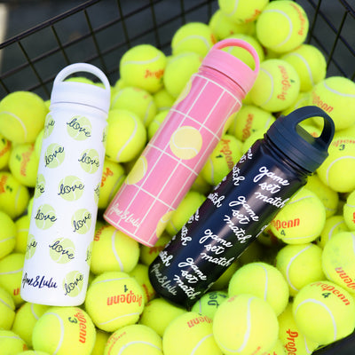 Basket of tennis balls holding tennis balls and three water bottles. Bottles are white with tennis balls, coral with yellow tennis balls, and navy bottle with game set match text.